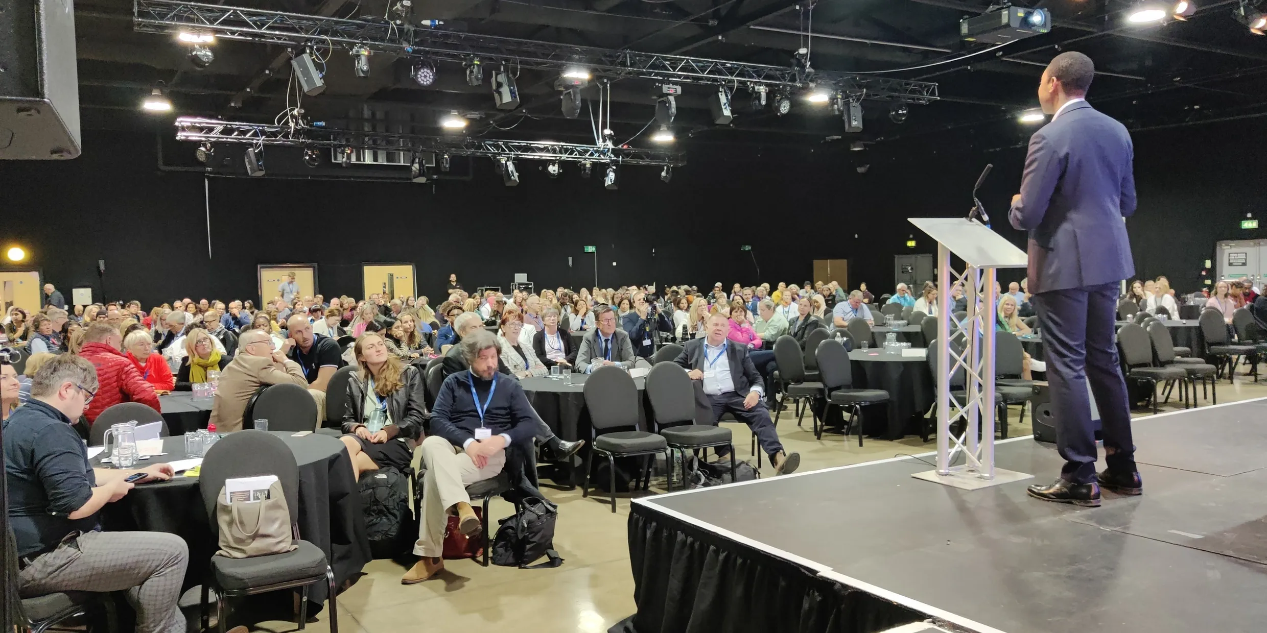 Speaker on stage presenting to a large in-person audience.